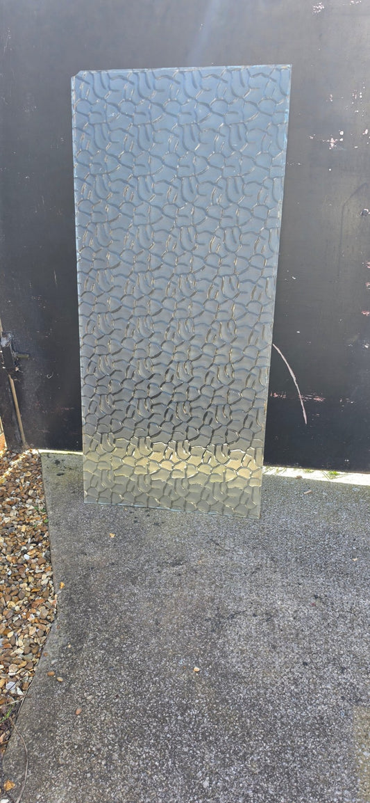 Rectangular sheet of textured glass leaning against a black metal gate on a concrete and gravel path.