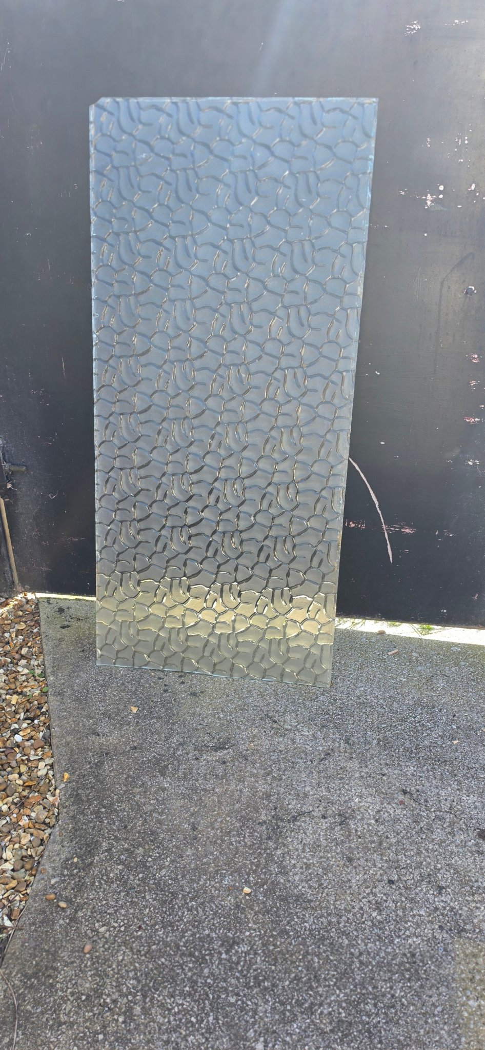 Rectangular sheet of textured glass leaning against a black metal gate on a concrete and gravel path.